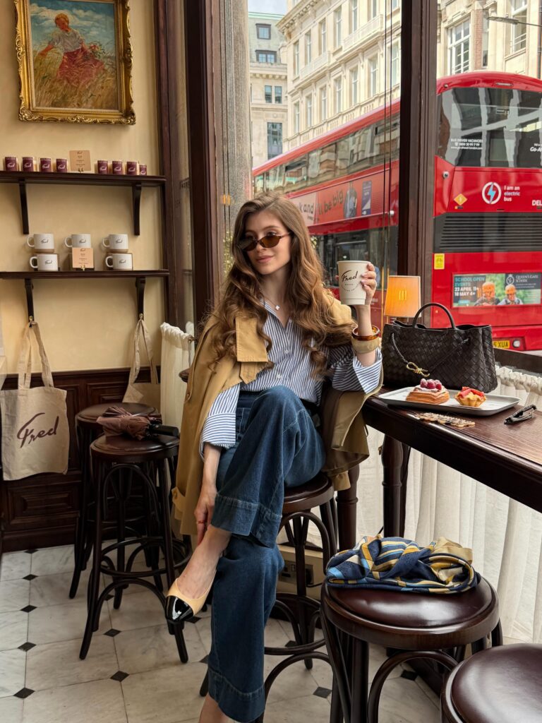 Tan trench coat over a blue-and-white striped shirt with jeans and two-tone slingback flats at a London café