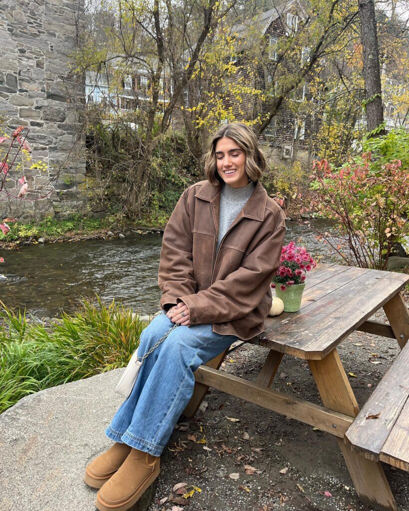 Oversized brown leather jacket over a grey knit with jeans and chestnut UGG mini boots beside a creek