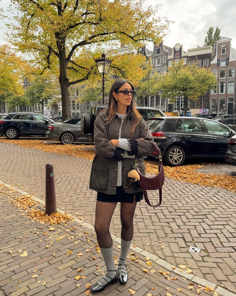 Waxed olive field jacket over a grey cardigan with a black mini skirt, tights, grey socks and Mary Jane flats among fallen leaves