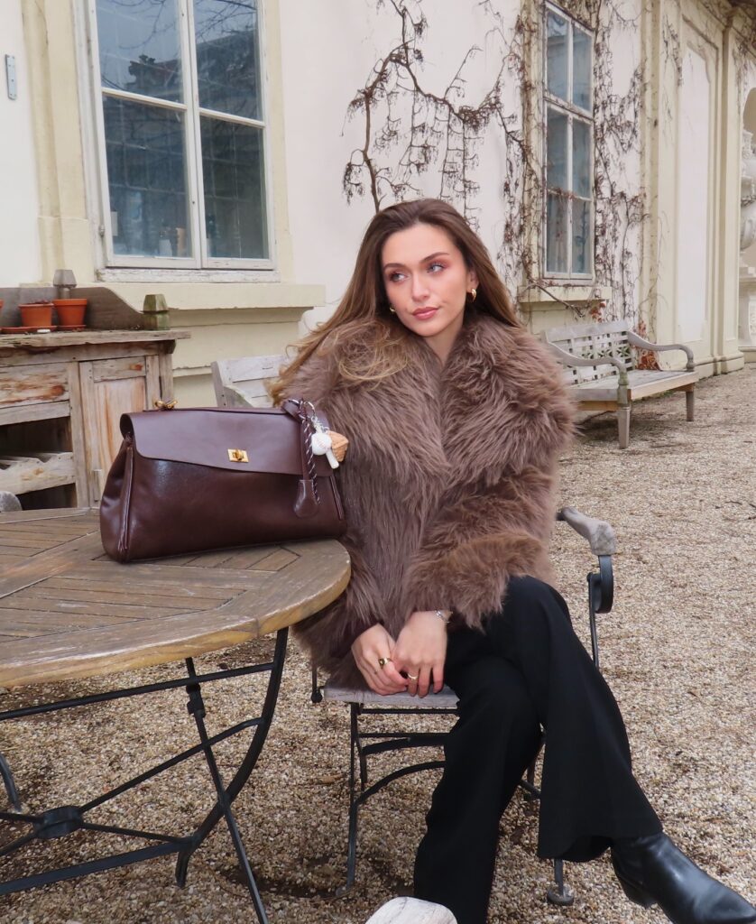 Taupe faux-fur jacket with black trousers, black boots and a brown leather bag in a courtyard