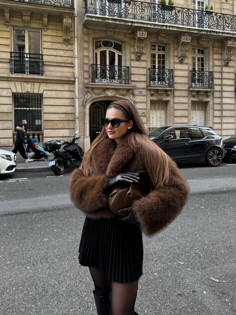 Brown fur jacket over a black pleated mini skirt with tights, leather gloves and a headband on a Paris street