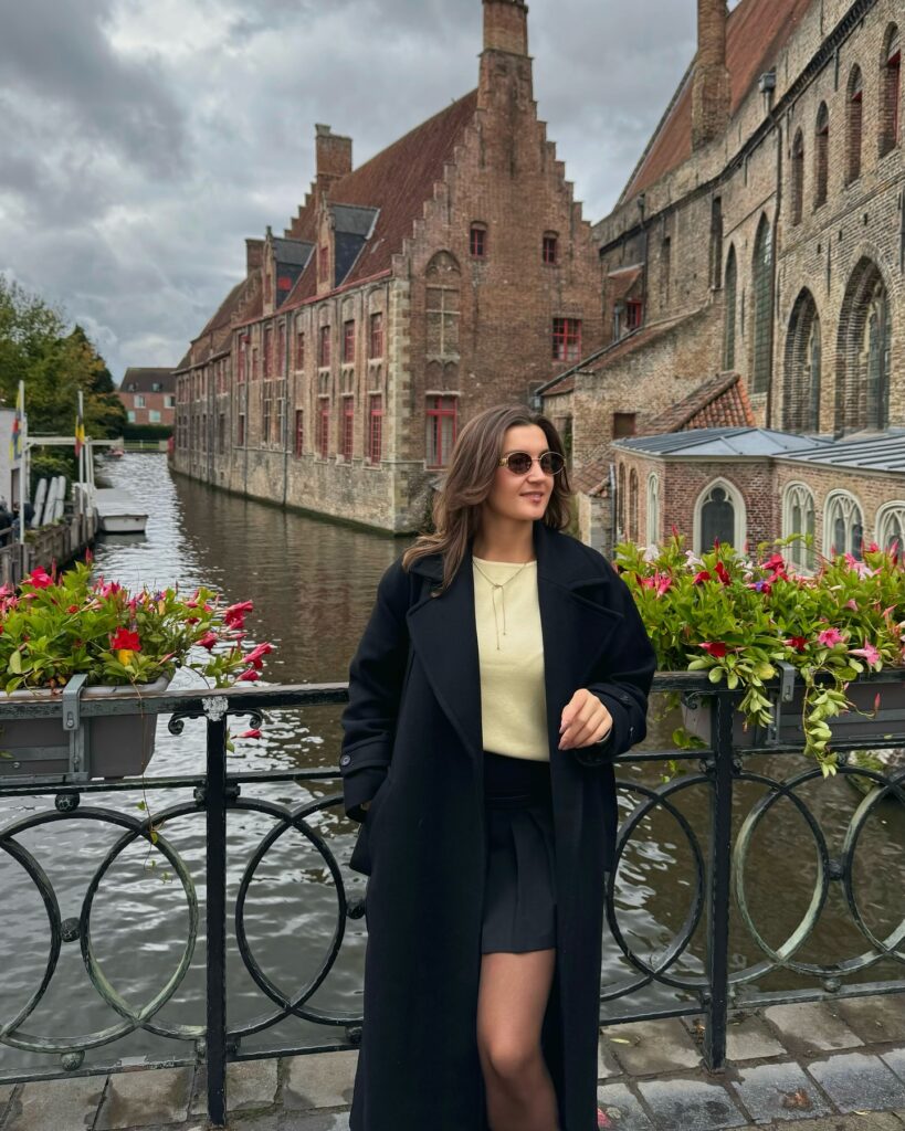 Black long wool coat over a pale yellow knit with a black skirt and tights by a Bruges canal