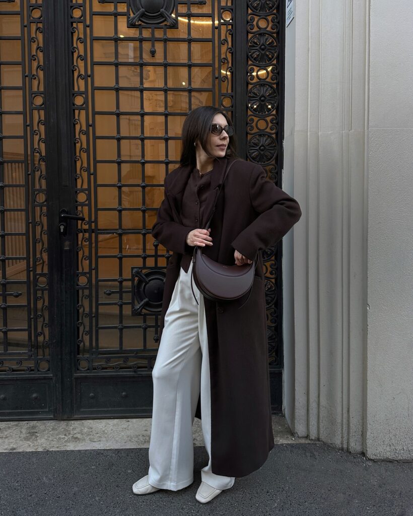 Dark brown long wool coat over a brown cardigan with white wide-leg trousers, white mules and a brown bag in Paris