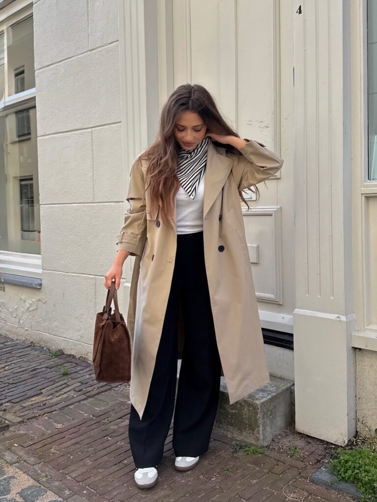 Beige trench coat with a white tee, striped silk scarf, black wide-leg trousers, white sneakers and a suede bucket bag in Amsterdam