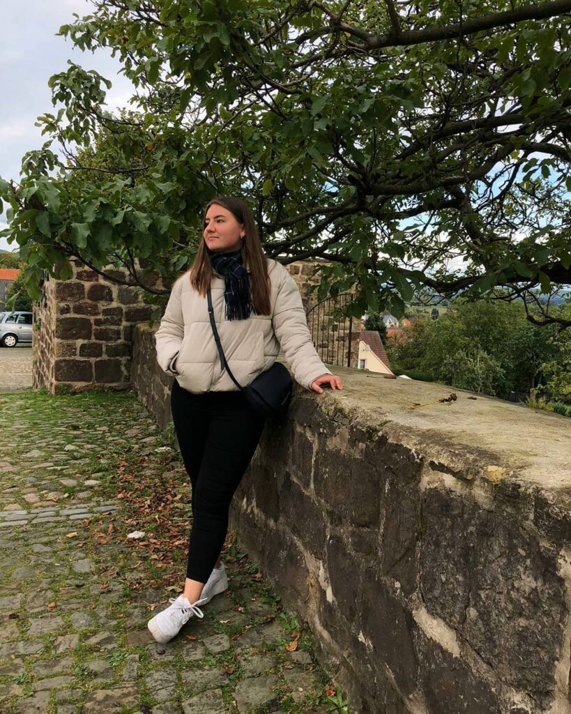 Cream puffer jacket with a navy scarf, black jeans and white sneakers on a castle wall