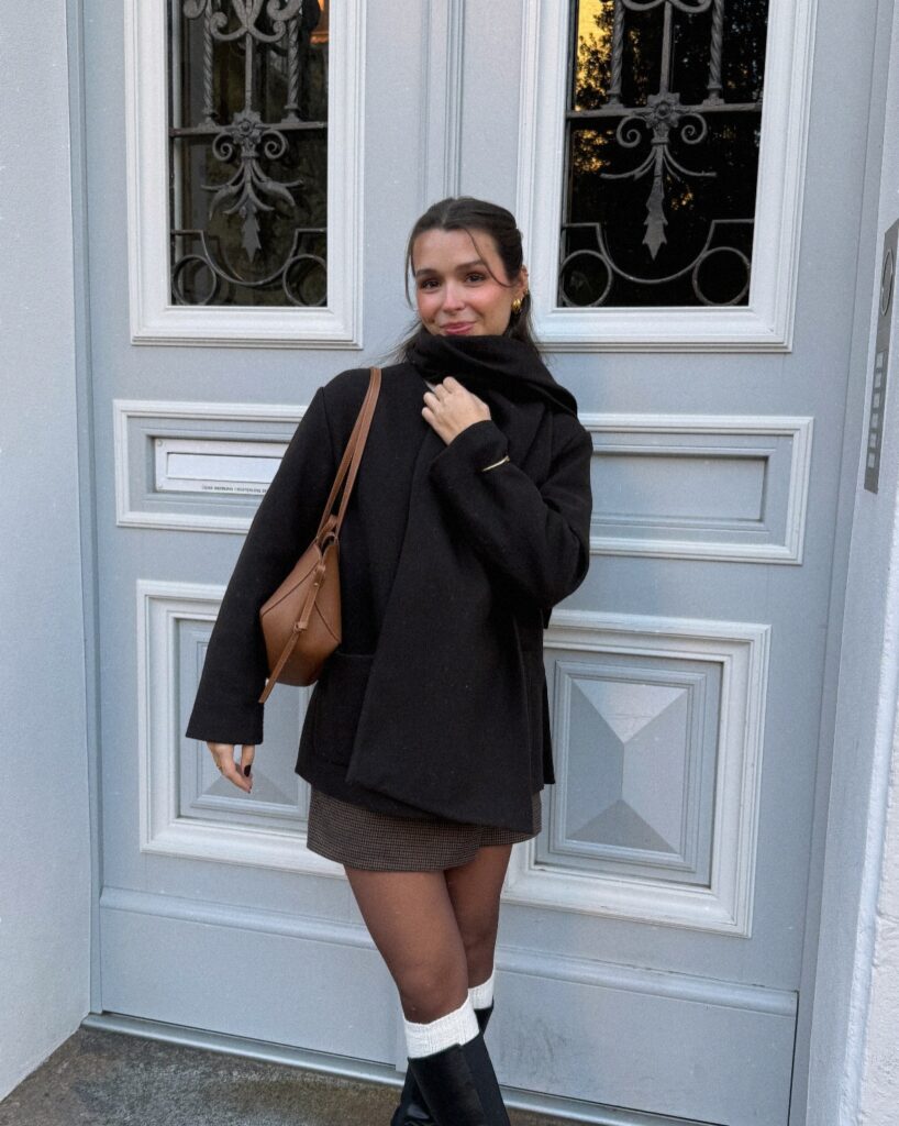 Black wool coat with a scarf, brown plaid mini skirt, white socks and black boots in front of a blue door