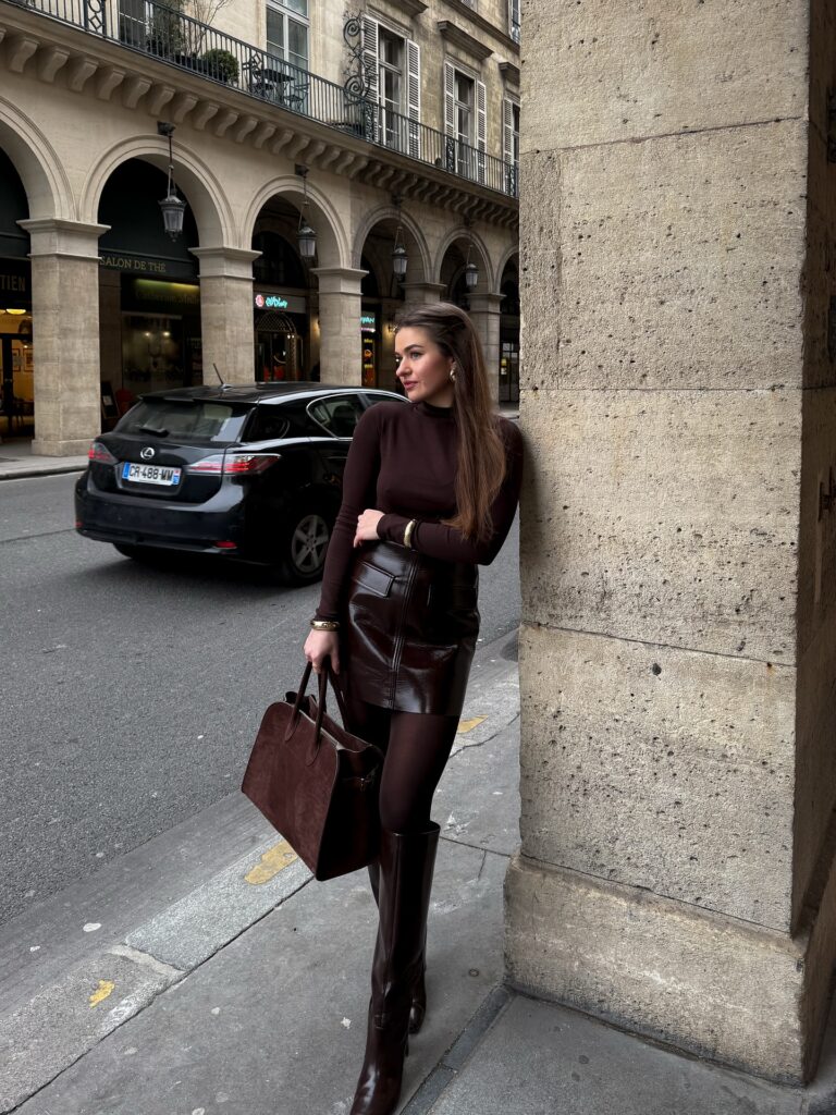 Brown turtleneck with a brown patent mini skirt, brown tights, brown knee-high boots and a suede tote under Paris arcades
