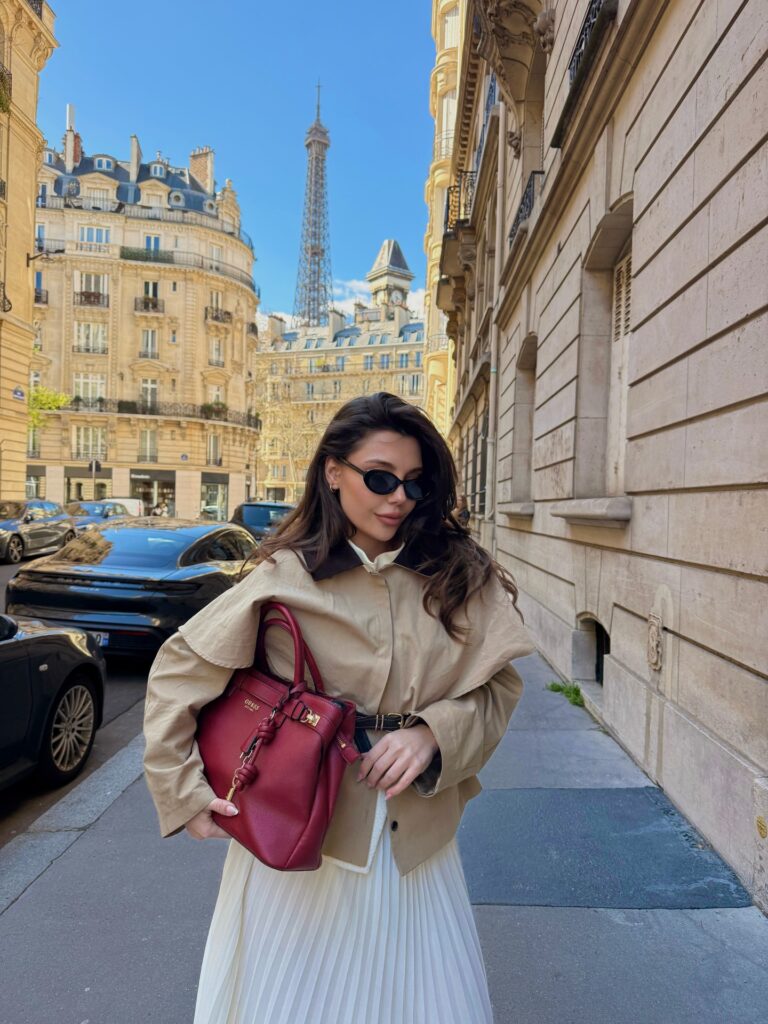 Cropped tan trench jacket over a cream pleated maxi skirt with a red top-handle bag, Eiffel Tower in the background
