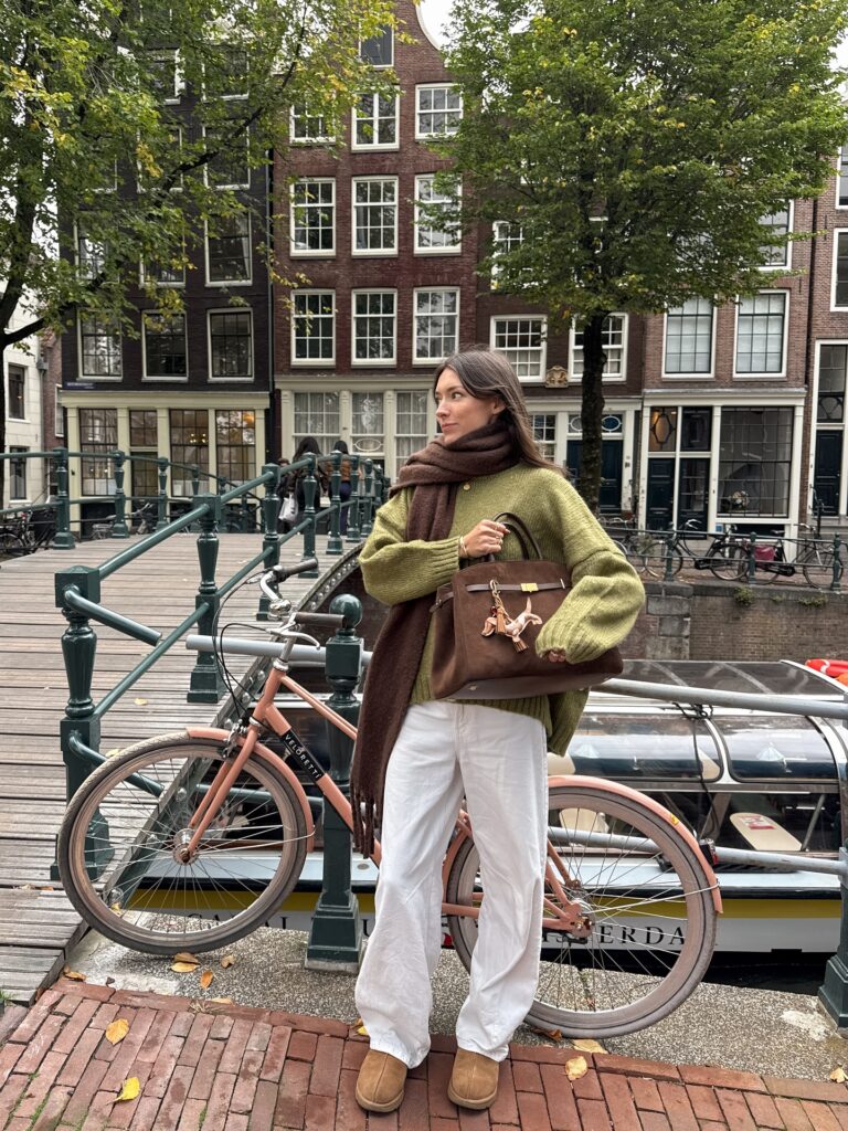 Olive green knit sweater with a brown scarf, white wide-leg jeans and chestnut UGG mini boots by an Amsterdam canal