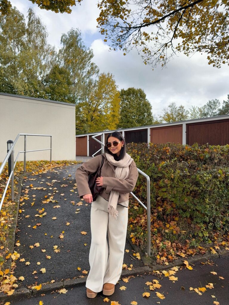Taupe cropped jacket with a scarf, cream knit wide-leg trousers and platform UGG boots among autumn leaves