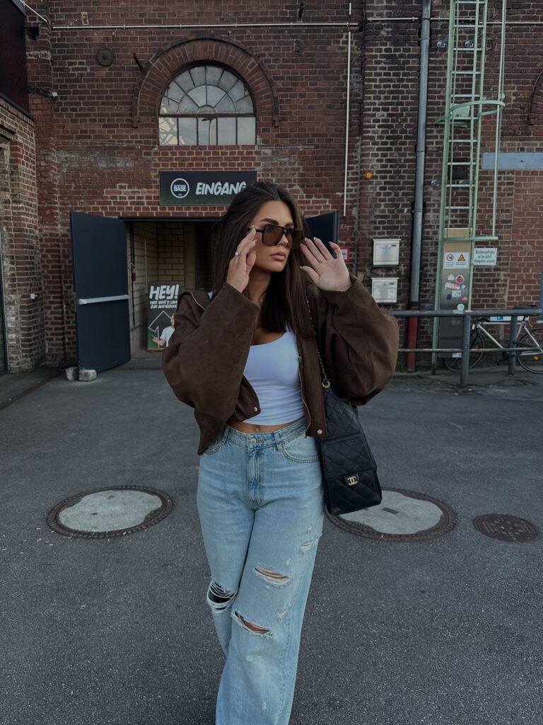 Woman in a cropped brown suede jacket, white tank top and light ripped wide-leg jeans with a black quilted bag, standing on a city street