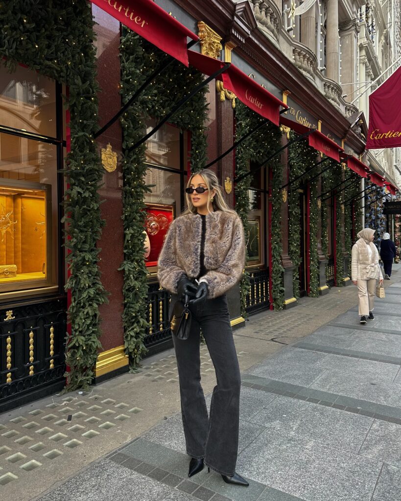 Woman in a taupe faux fur jacket, black top, charcoal wide-leg jeans and pointed black boots in front of a festively decorated Cartier storefront