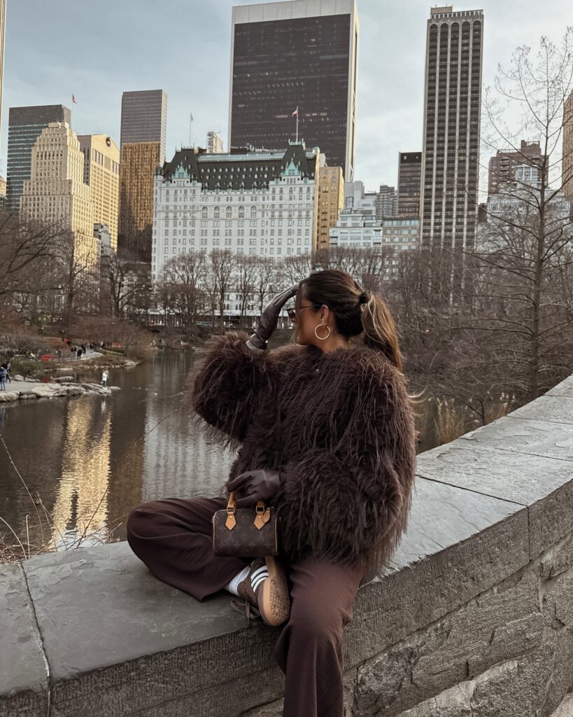 Woman seated on a stone ledge in a shaggy brown faux fur jacket, brown trousers, leather gloves and brown Adidas Samba sneakers, with the Plaza Hotel and Central Park behind her