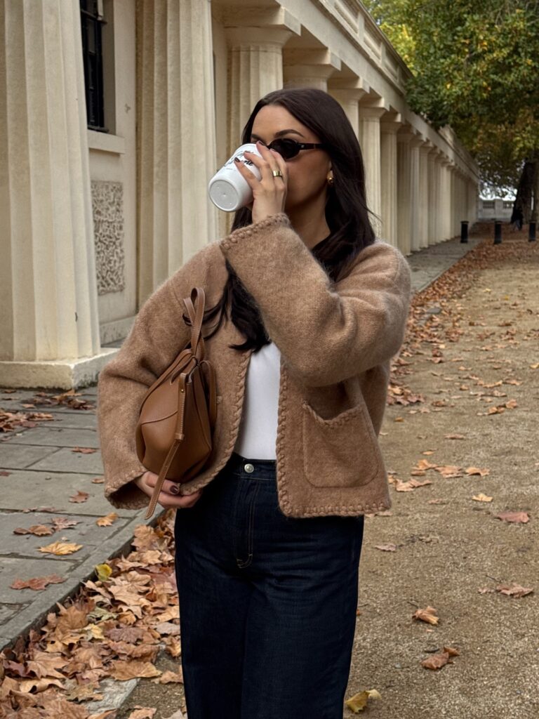 Woman sipping coffee in a camel knit cardigan over a white top with dark jeans, holding a tan leather bag, among autumn leaves and columns