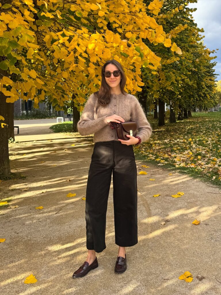 Woman in a fuzzy mauve-taupe mohair cardigan, black wide-leg trousers and burgundy loafers, holding a burgundy bag under a golden autumn tree