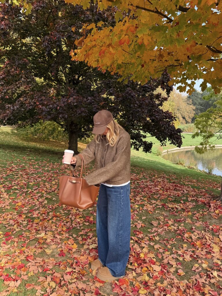 Blonde woman in a brown baseball cap, brown knit cardigan, white tee and baggy blue jeans, holding a coffee and tan tote among fallen autumn leaves