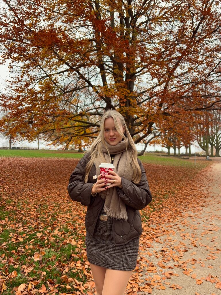 Woman by a lake in a brown quilted jacket, taupe scarf and grey plaid mini skirt, holding a red coffee cup among autumn trees