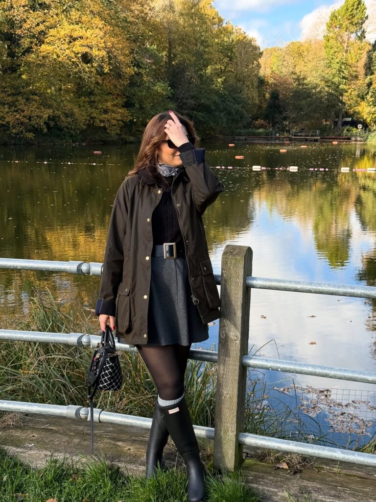Woman beside a lake in an olive waxed field jacket over a black cable knit, grey skirt, tights and tall black Hunter wellington boots