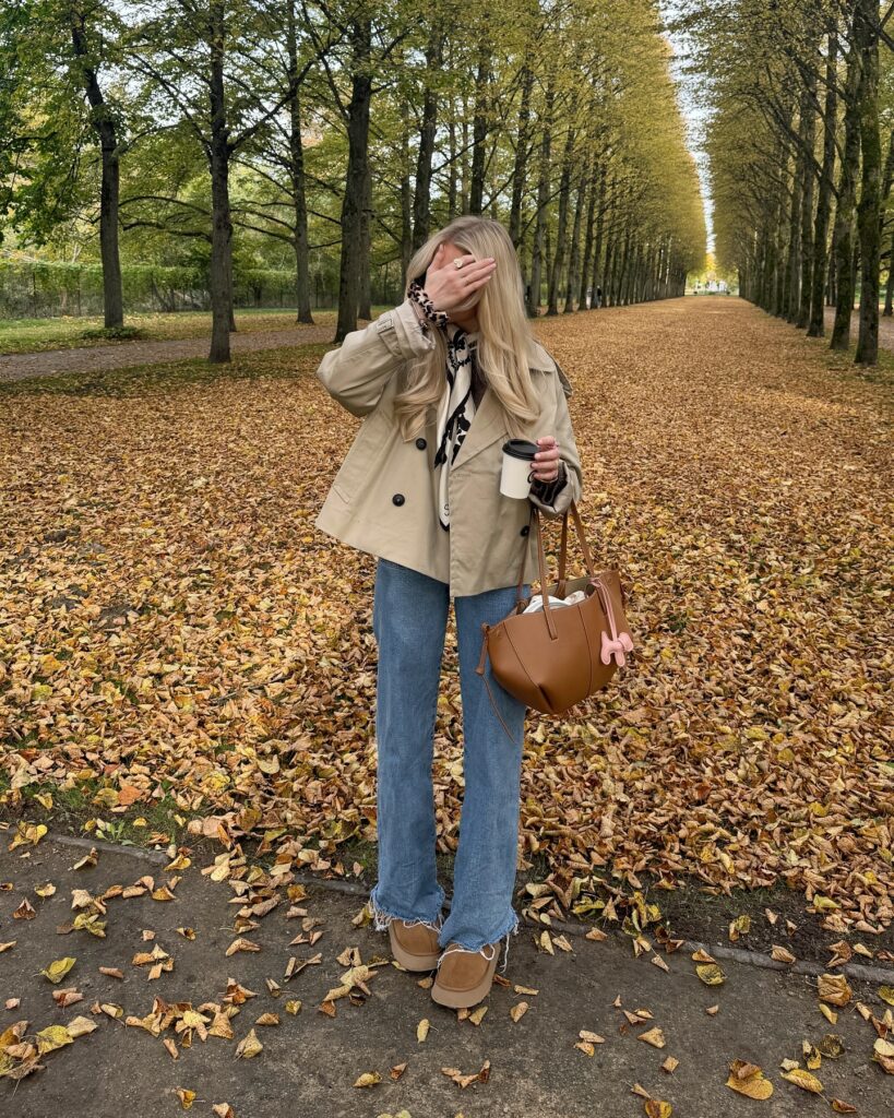 Blonde woman in a short khaki trench jacket with a leopard print scarf, blue flared jeans and tan slippers, holding a coffee and brown tote on a leaf-covered avenue