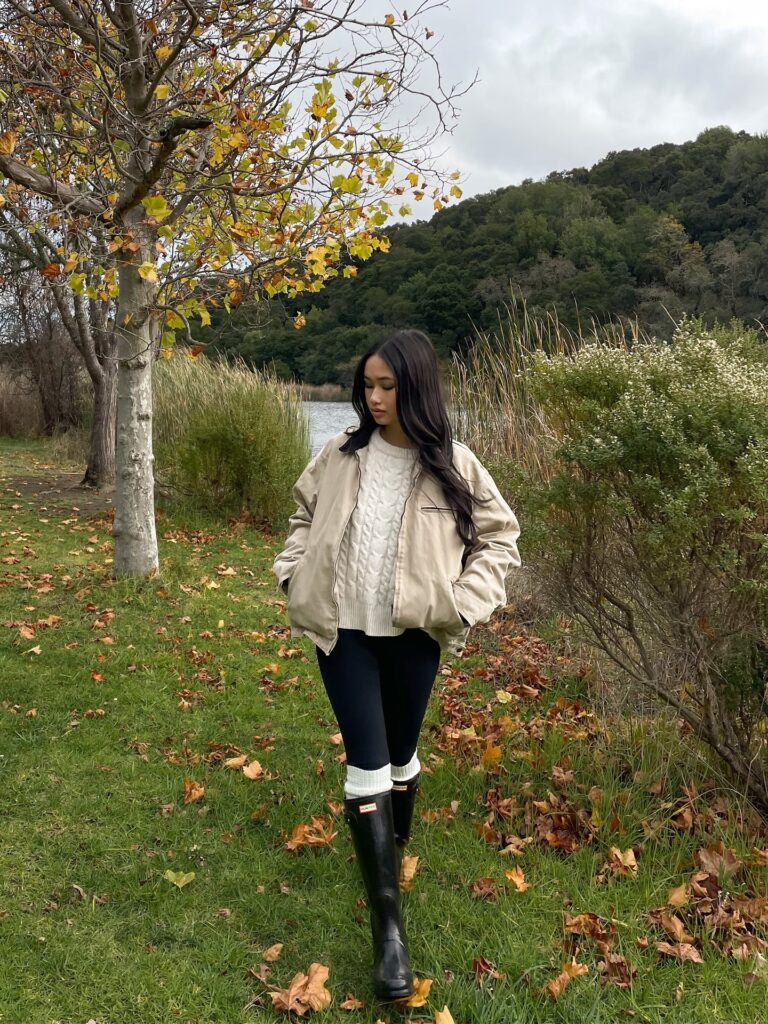 Woman walking by a lake in a cream bomber jacket over a cream cable knit sweater with black leggings, white socks and tall black Hunter boots