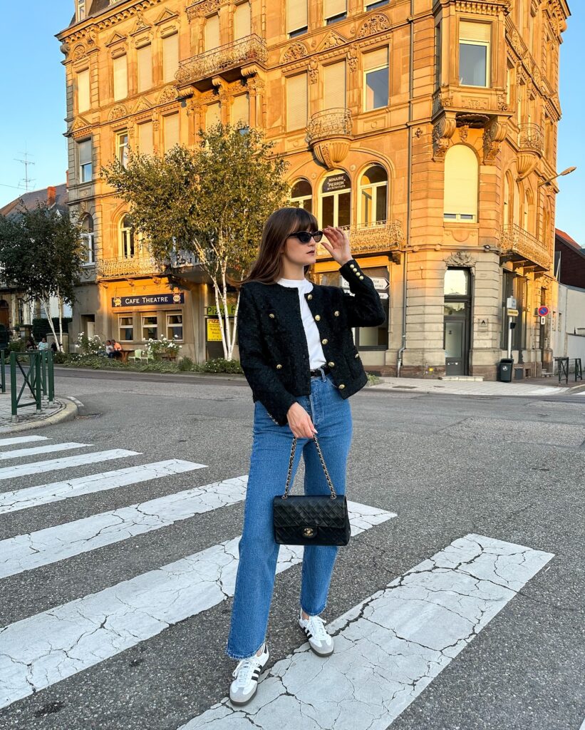 Woman crossing a street in a black tweed jacket with gold buttons, white tee and blue jeans, holding a black quilted bag and wearing Adidas Samba sneakers
