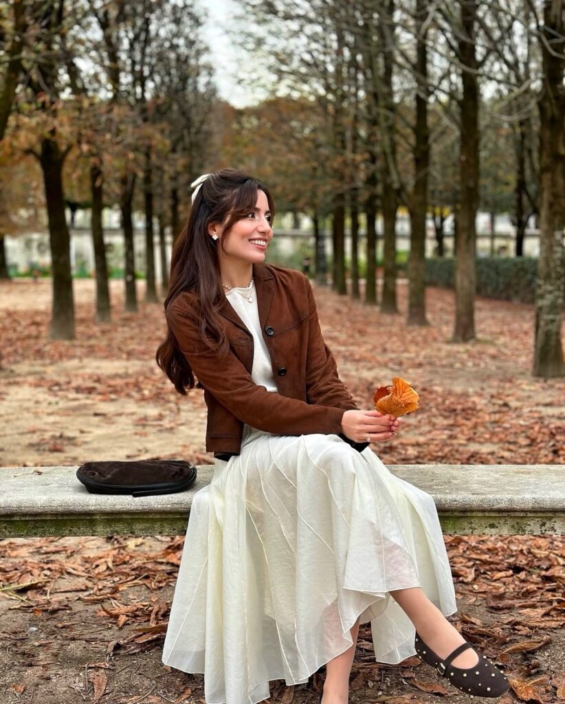 Smiling woman sitting on a stone bench in a cropped brown suede jacket over a cream top and floaty cream maxi skirt with suede Mary Jane flats in an autumn park