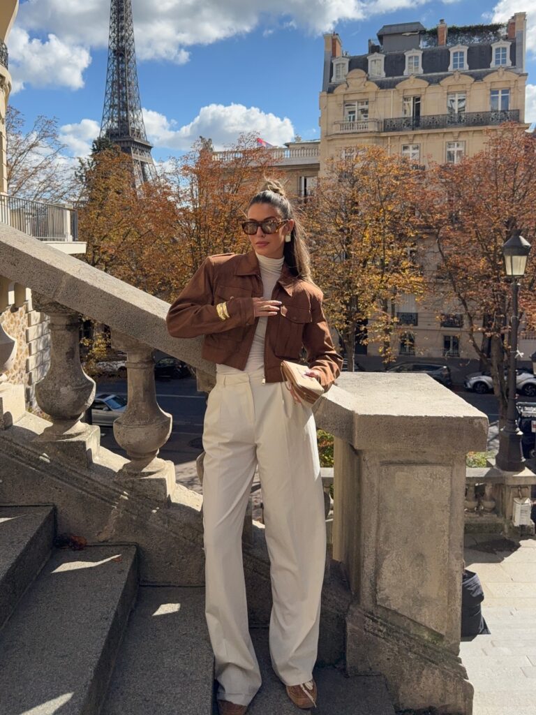 Woman in a rust brown suede jacket, cream turtleneck and cream wide-leg trousers on a Paris staircase with the Eiffel Tower behind her