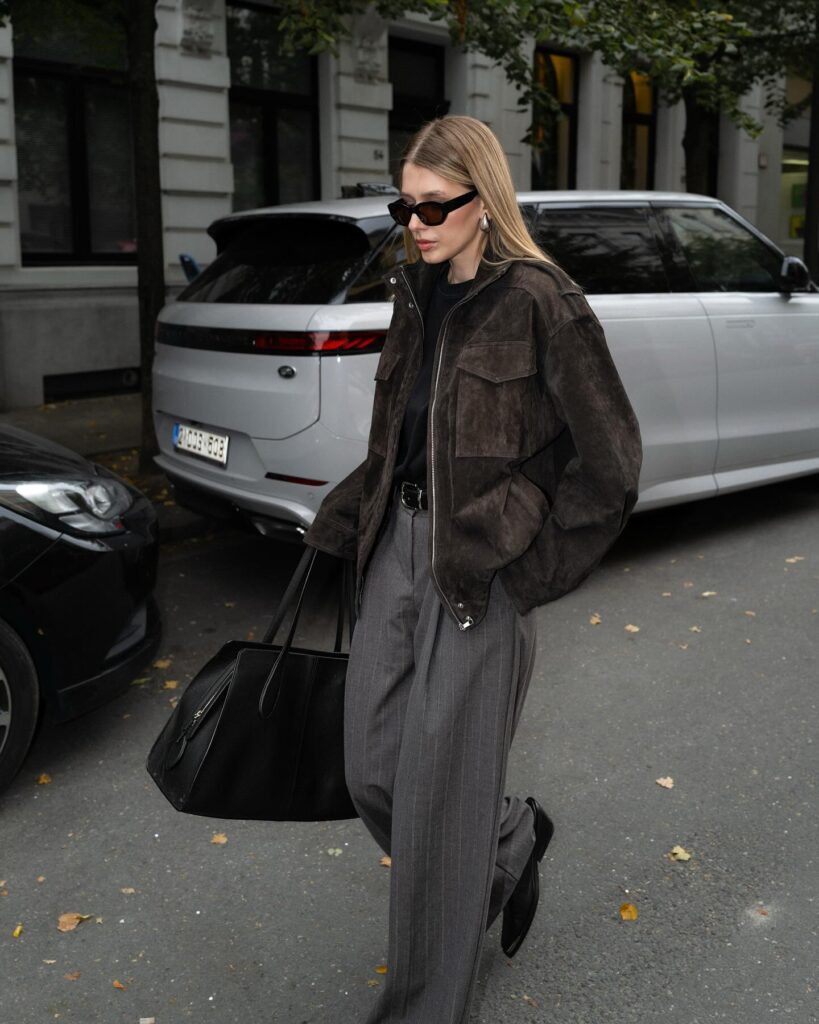 Woman walking in a dark brown suede zip jacket over a black tee with grey pinstripe wide-leg trousers, carrying a large black tote