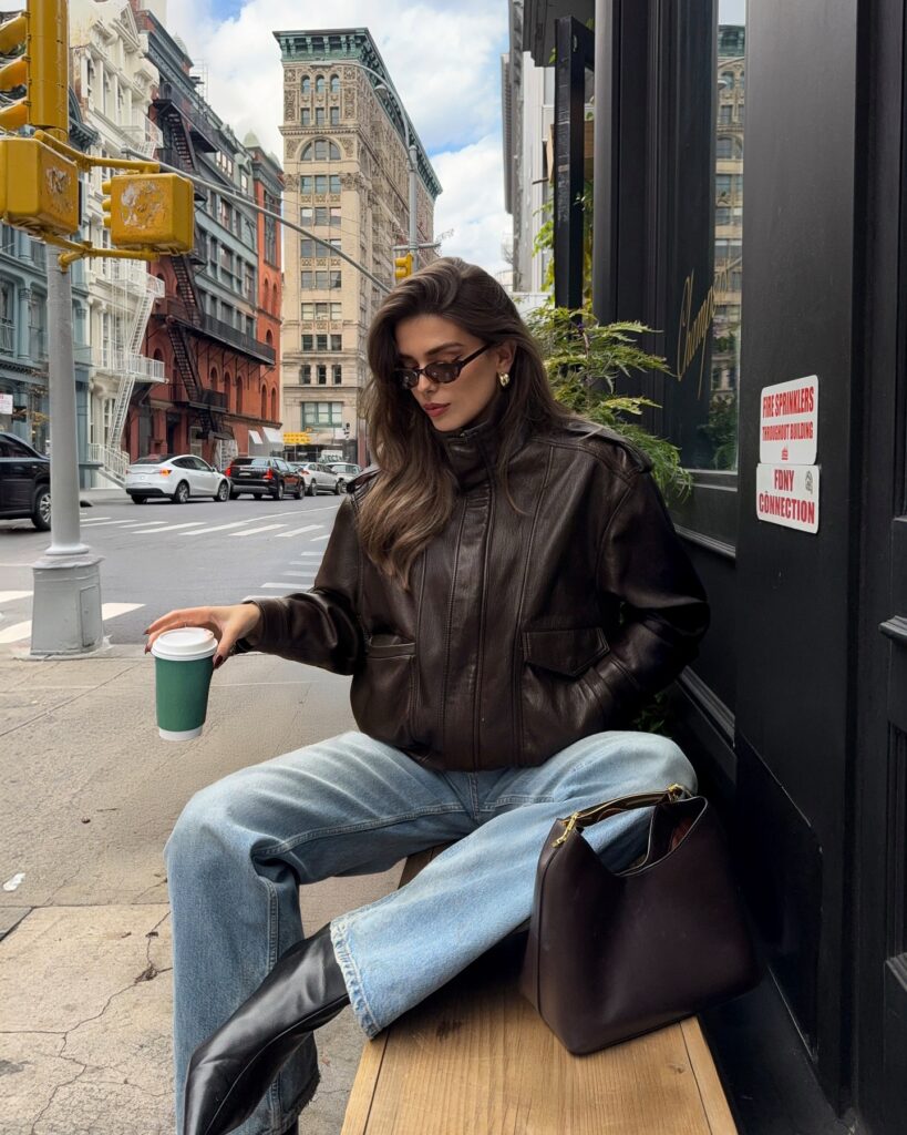 Woman sitting on a bench in a worn brown leather jacket, light faded wide-leg jeans and black boots, holding a coffee cup on a New York street