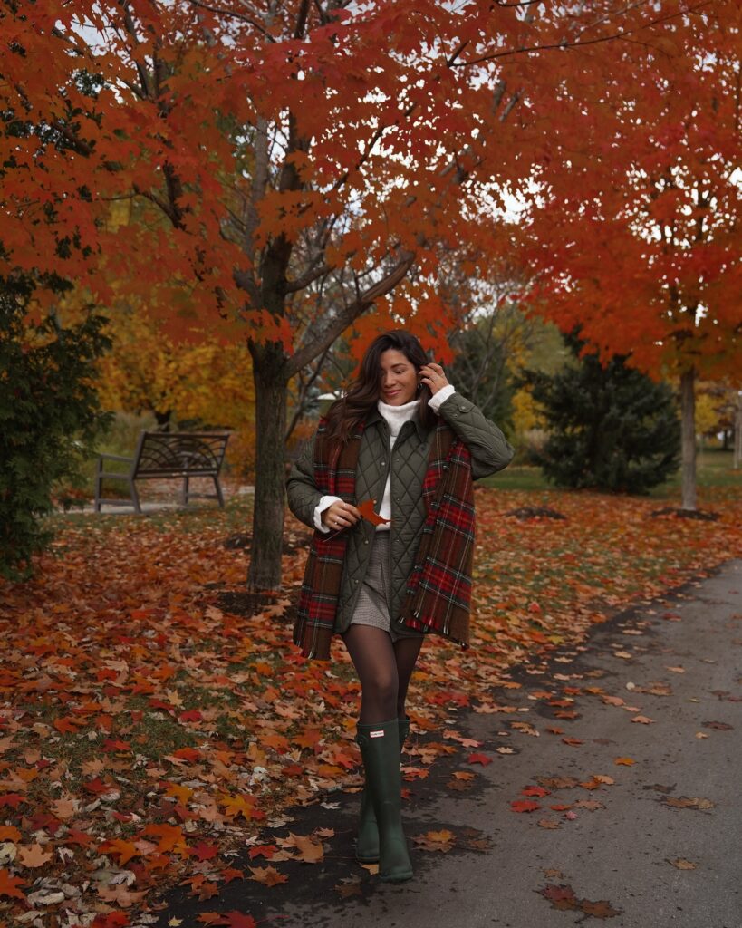 Olive quilted jacket with a red tartan scarf, white turtleneck, tweed mini skirt, tights and green wellington boots on a leaf-covered path