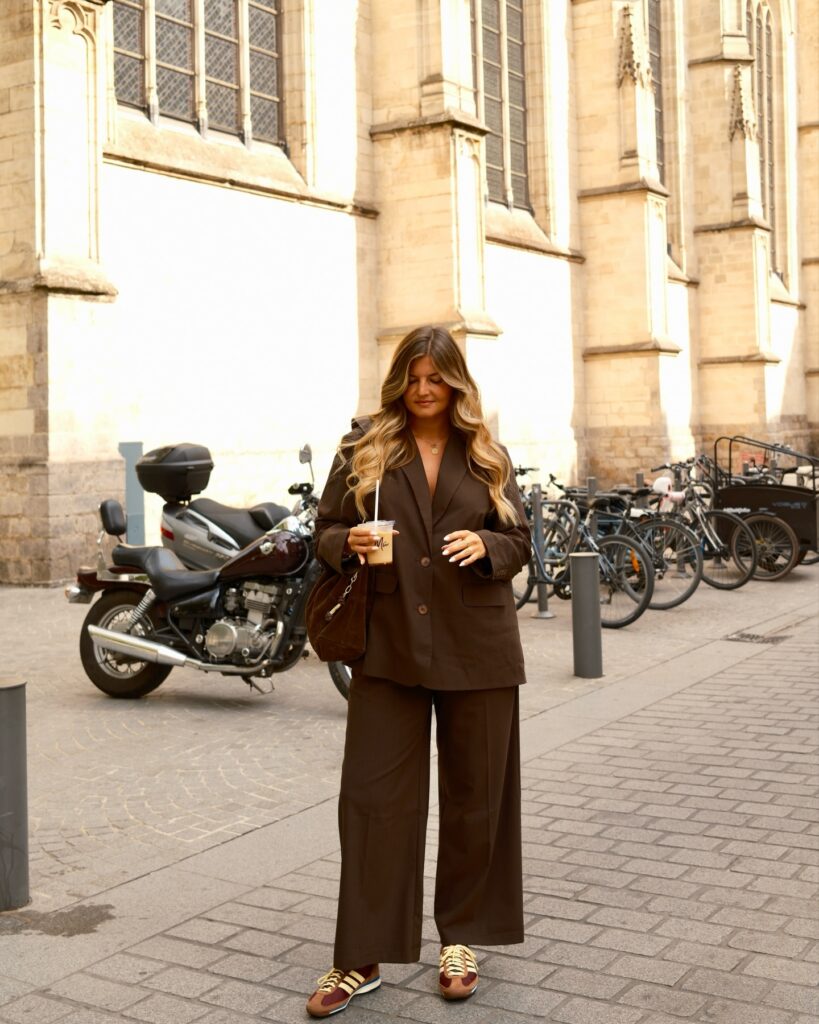Chocolate-brown oversized blazer and matching wide-leg trousers with burgundy-and-tan sneakers, holding an iced coffee
