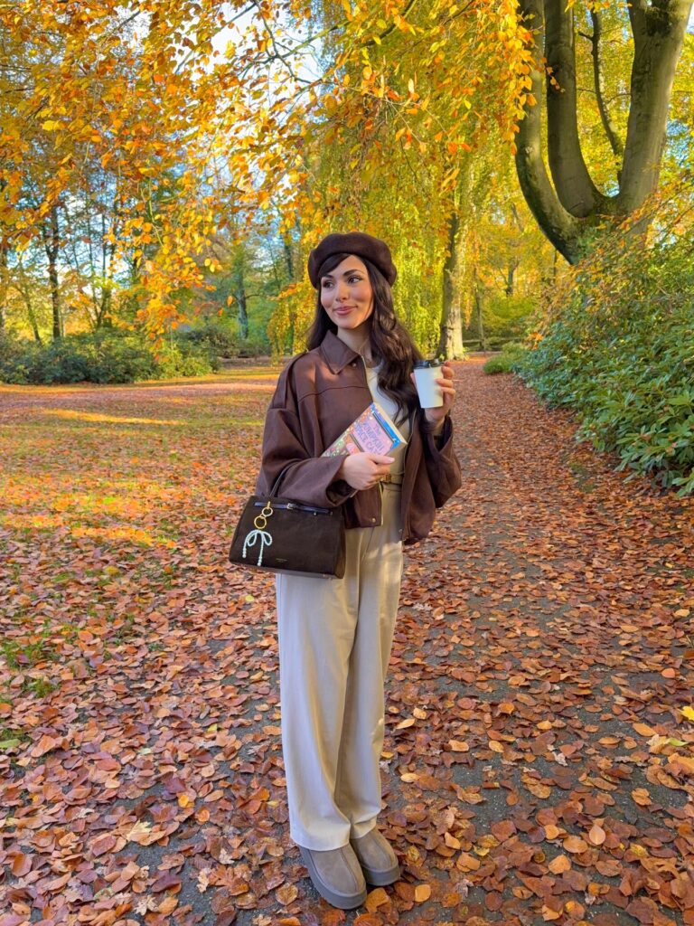 Brown jacket over a cream wide-leg co-ord with a brown beret and platform boots, holding a coffee and a book