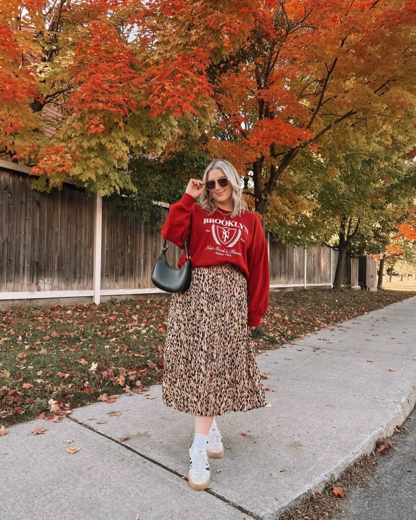 Red Brooklyn graphic sweatshirt tucked into a leopard-print pleated midi skirt with Adidas sneakers and a black shoulder bag