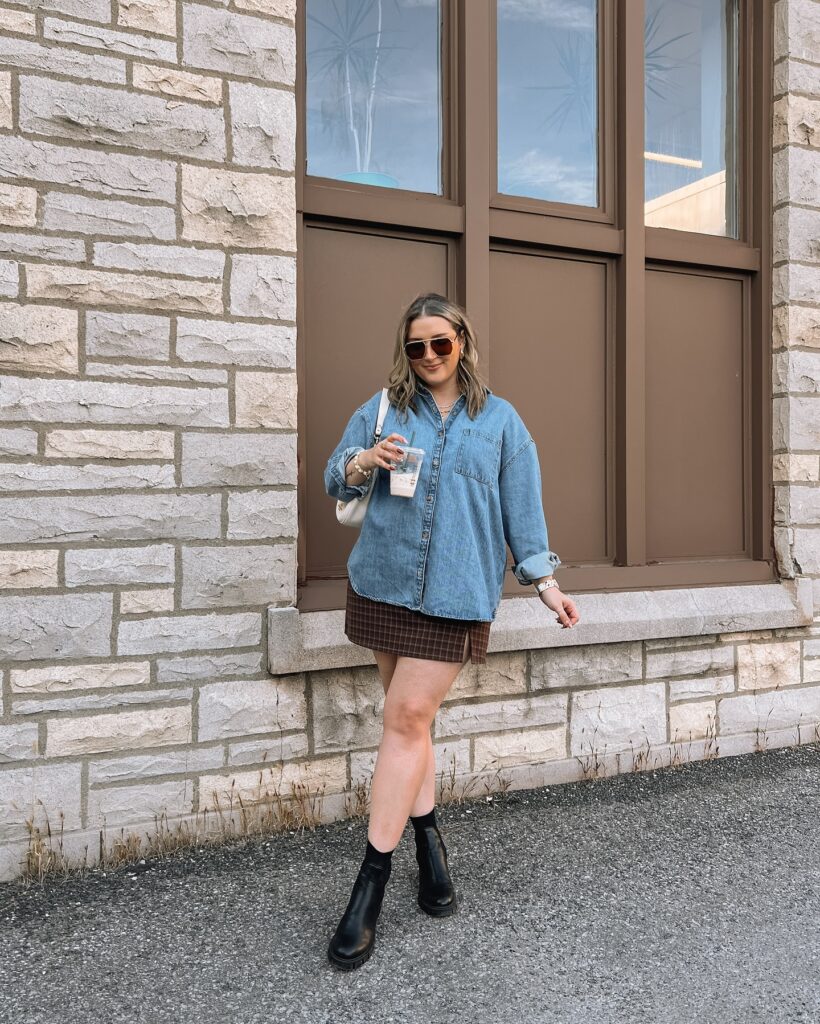 Oversized denim shirt with a brown plaid mini skirt and black chunky ankle boots, holding an iced coffee