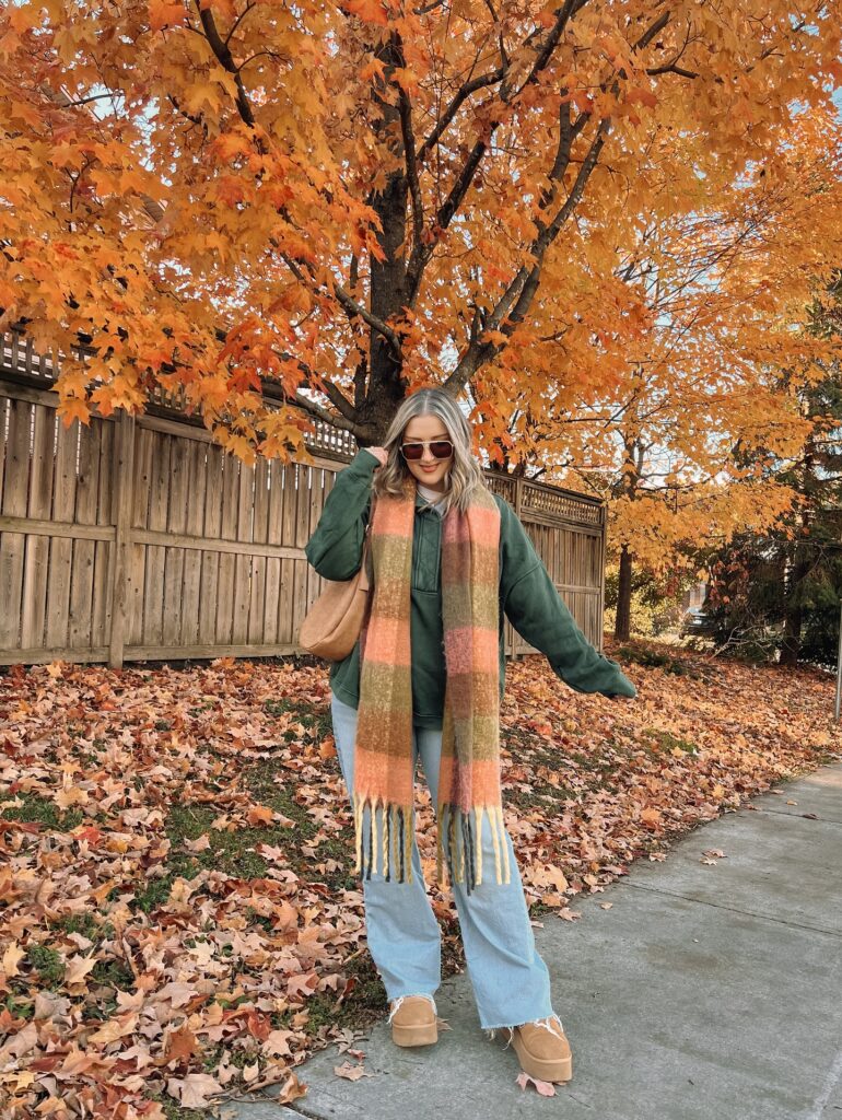Green hoodie with a plaid fringed scarf, light-wash wide-leg jeans and tan platform boots under an orange autumn tree
