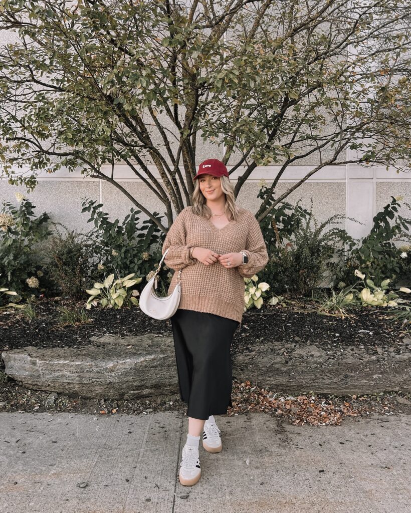 Camel V-neck sweater layered over a black satin slip skirt with white Adidas sneakers and a red cap