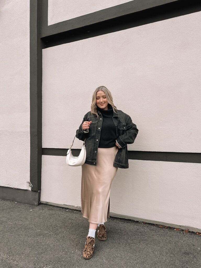 Washed-black oversized denim jacket over a black turtleneck with a champagne satin slip skirt and leopard-print sneakers