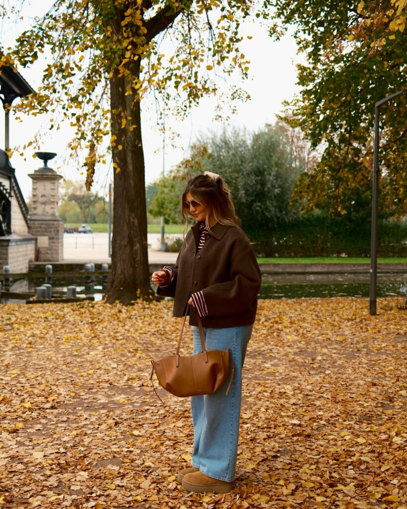 Brown wool jacket over a striped top with light-wash wide-leg jeans, tan platform boots and a tan leather tote in a leaf-covered park