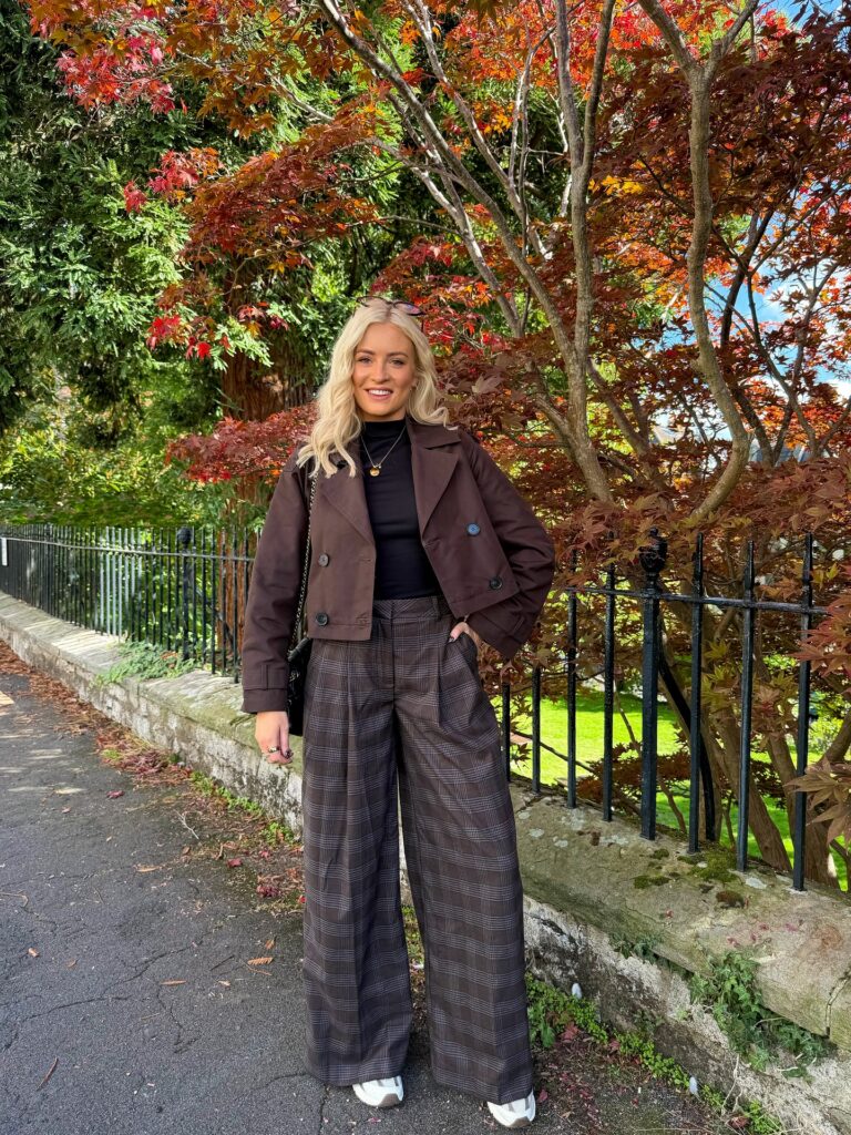 Brown cropped trench coat over a black turtleneck with brown plaid wide-leg trousers and white sneakers