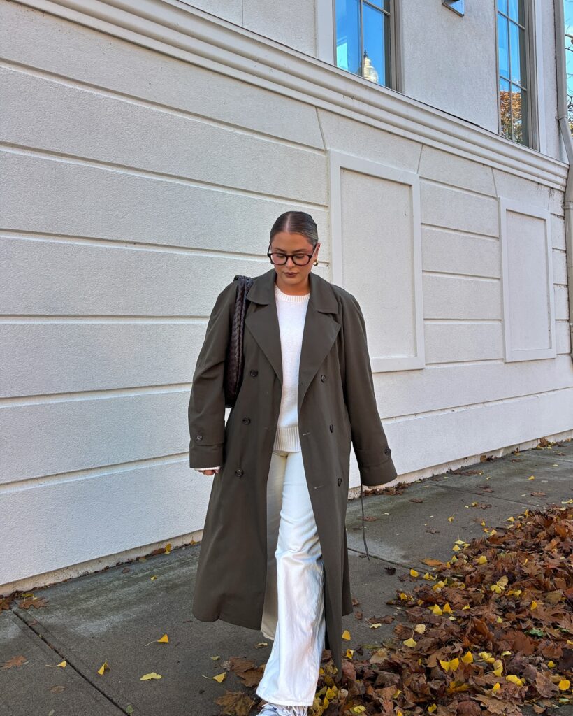 Olive double-breasted trench coat over a white knit and white trousers with sneakers