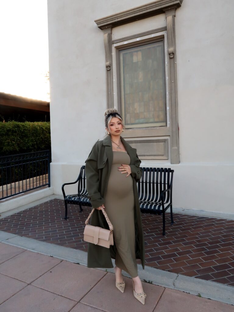 Pregnant woman in a long olive trench coat layered over an olive bodycon maxi dress with nude heels and a tan top-handle bag