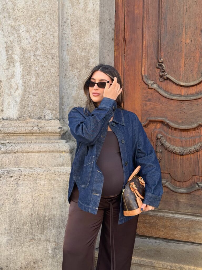 Pregnant woman in an indigo denim chore jacket over a fitted brown bodysuit and brown wide-leg trousers holding a monogram bag