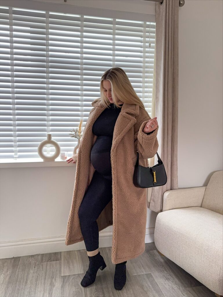 Pregnant woman in a long camel teddy coat over a black activewear set with black ankle boots and a black shoulder bag