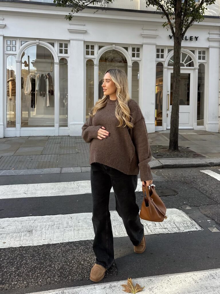 Pregnant woman in a brown crewneck sweater with black wide-leg jeans, tan UGG mini boots and a tan top-handle bag at a crosswalk