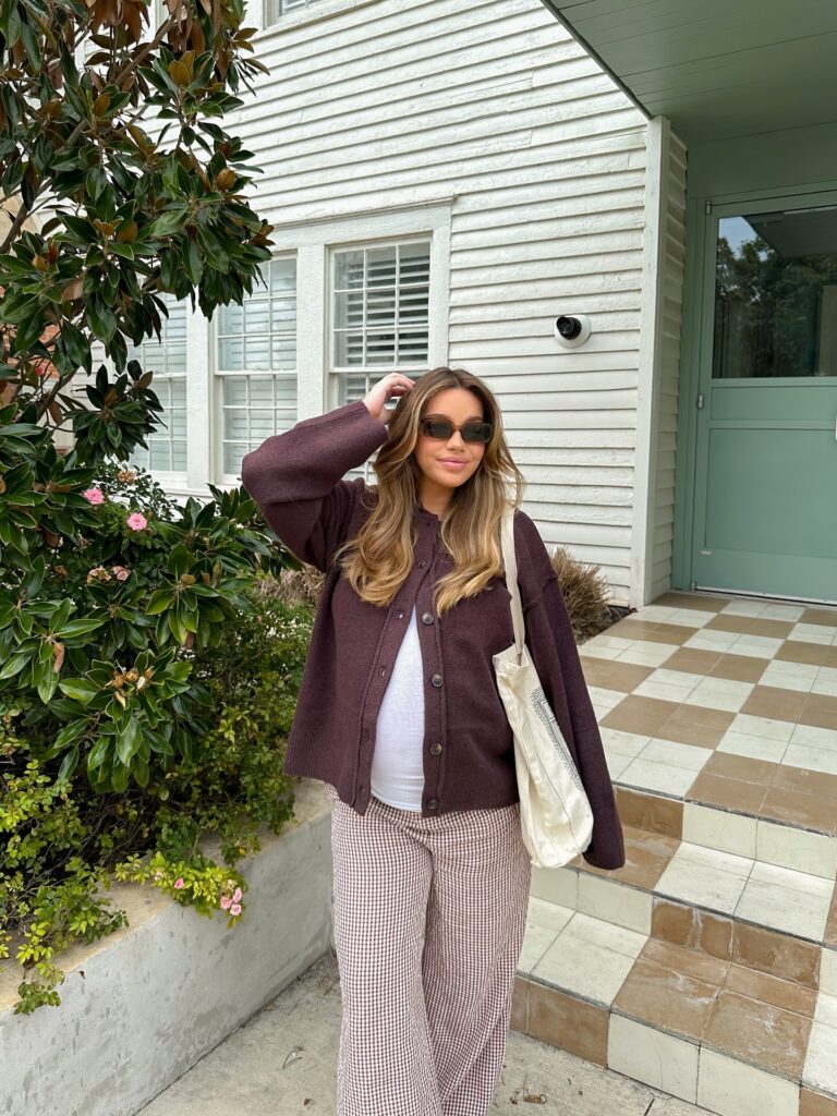 Pregnant woman in a chocolate button cardigan over a white tank with brown gingham wide-leg pants and a canvas tote