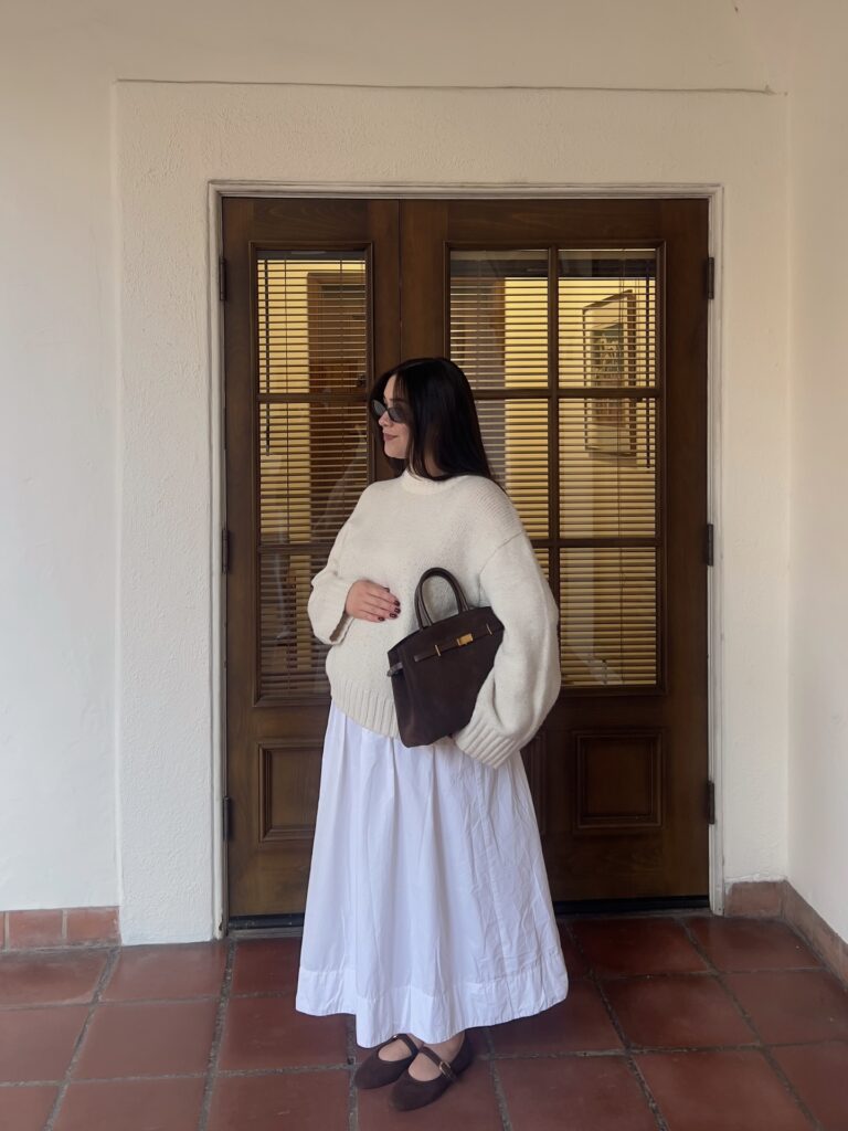 Pregnant woman in a cream chunky sweater over a white cotton maxi skirt with a brown suede top-handle bag and brown mary-jane flats