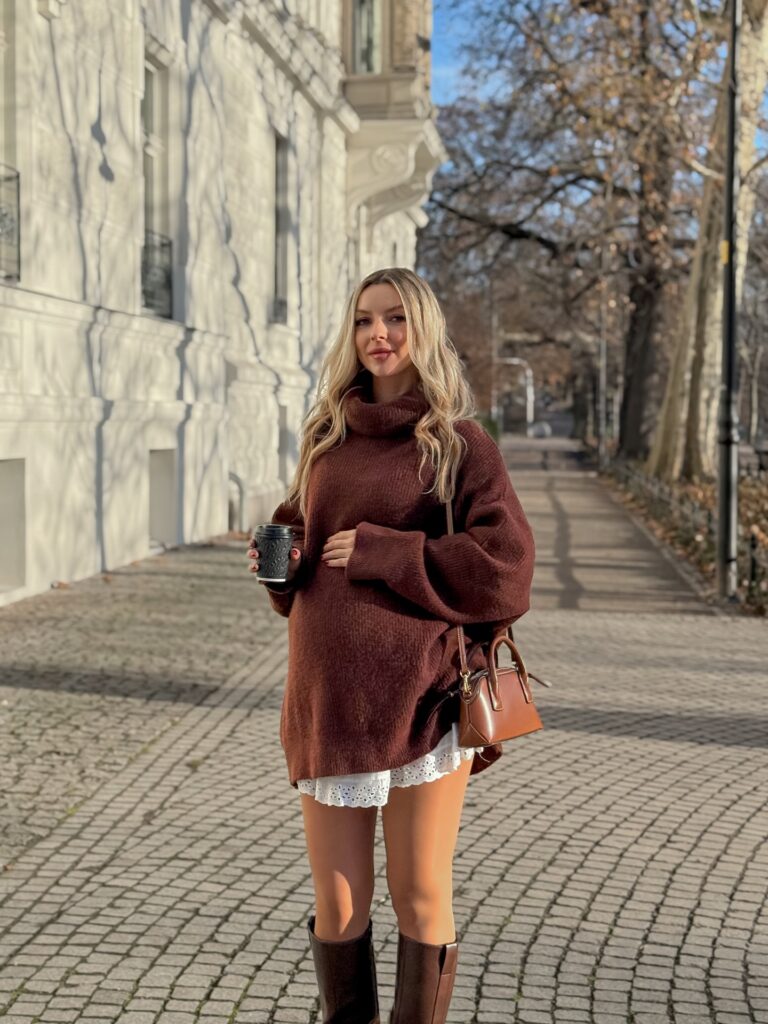 Pregnant woman in a brown oversized turtleneck sweater over a white broderie skirt with brown knee-high boots holding a coffee