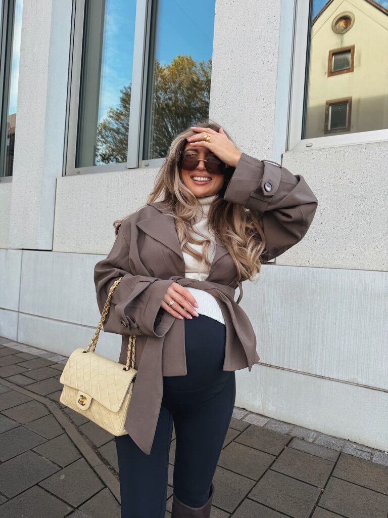 Pregnant woman in a taupe belted trench jacket, cream chunky turtleneck and navy leggings carrying a quilted cream flap bag