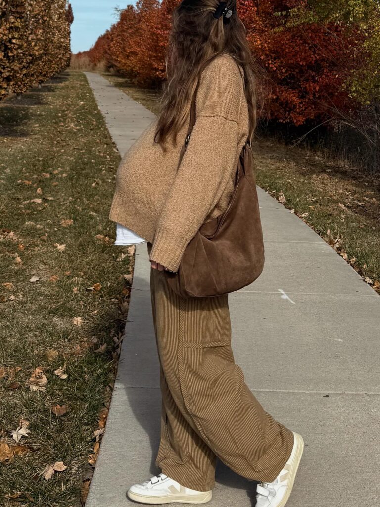 Pregnant woman in a camel sweater with brown corduroy wide-leg trousers, a suede shoulder bag and white Veja sneakers on an autumn path