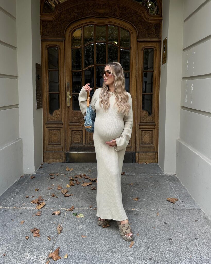 Pregnant woman in an oat ribbed knit maxi dress with platform slides and a small denim bag among fallen autumn leaves